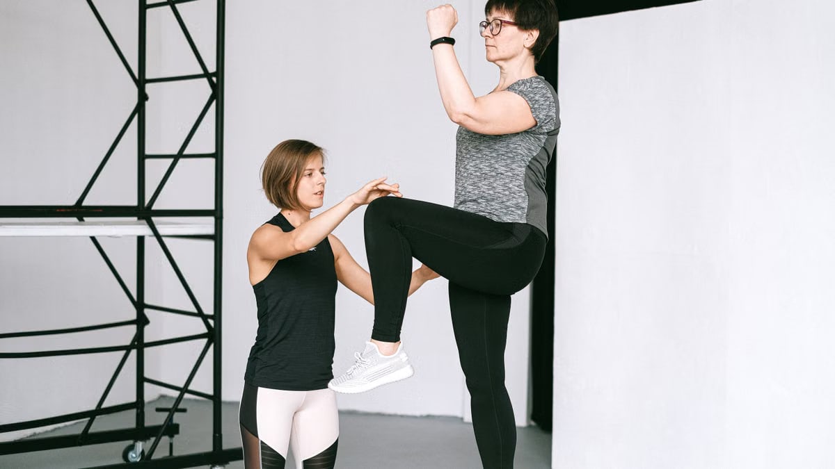 An elderly woman performs exercises with a fitness coach, focusing on a healthy lifestyle and wellness indoors.
